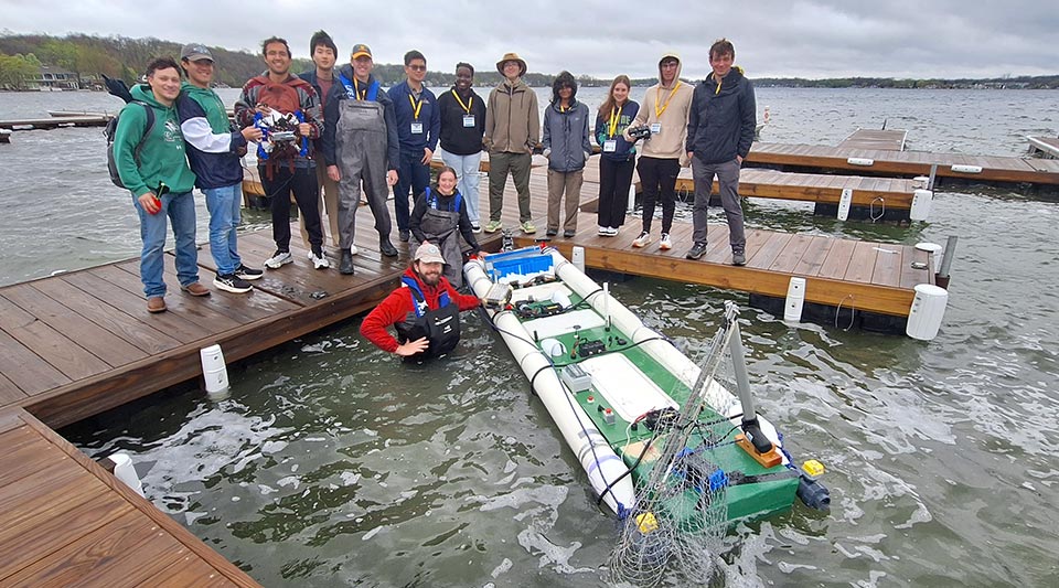 Twelve students stand on a dock while one female student sits with her feet in the water. Another student stands in the water and poses with the modular boat.